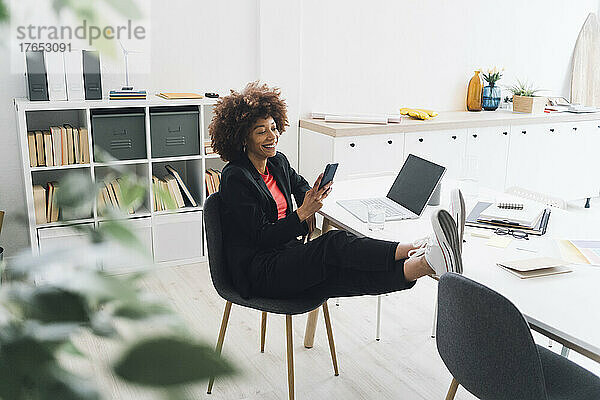 Lächelnde Geschäftsfrau  die mit erhobenen Füßen auf dem Schreibtisch im Büro sitzt und ihr Mobiltelefon benutzt