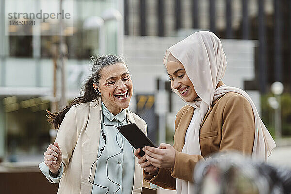 Glückliche Geschäftsfrau mit In-Ear-Kopfhörern genießt die gemeinsame Nutzung eines Tablet-PCs durch eine Kollegin