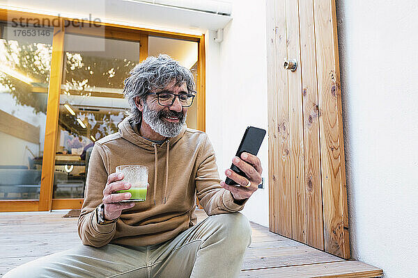 Lächelnder Mann mit einem Glas Smoothie sitzt auf der Terrasse