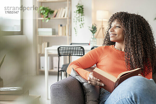 Glückliche junge Frau mit Buch sitzt zu Hause auf dem Sofa im Wohnzimmer