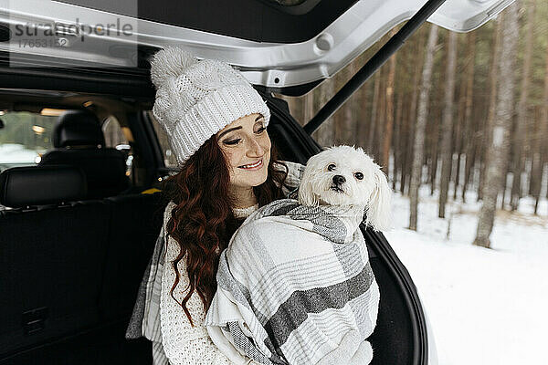 Lächelnde Frau mit süßem Hund  der in eine Decke gehüllt ist und im Kofferraum eines Autos sitzt