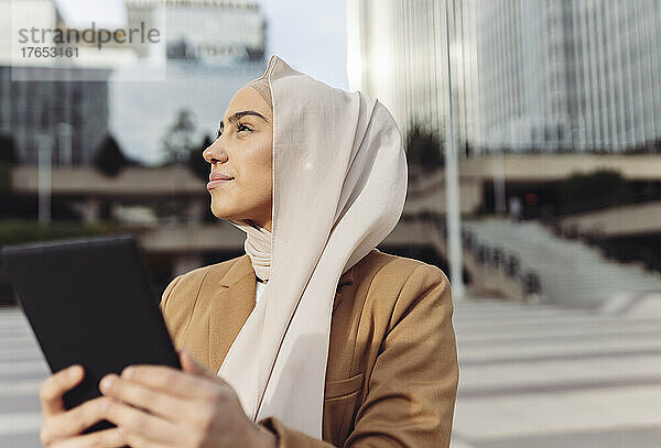 Junge Geschäftsfrau trägt Hijab und hält an einem sonnigen Tag einen Tablet-PC