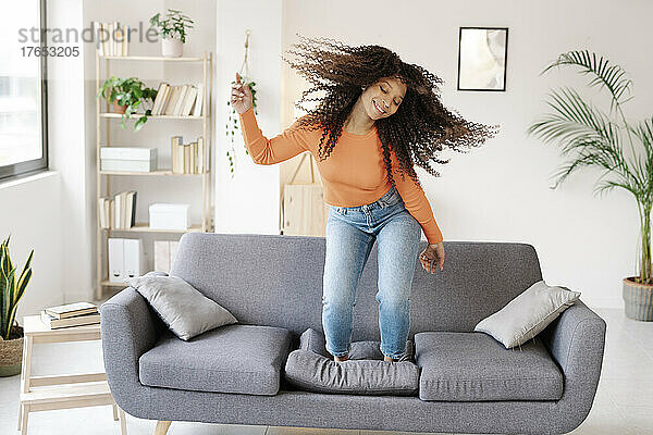 Lächelnde Frau mit zerzausten Haaren tanzt zu Hause auf dem Sofa
