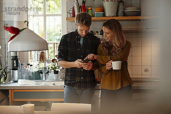 Frau hält Kaffeetasse in der Hand und zeigt auf Smartphone  das ihr Freund in der Küche benutzt