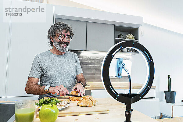 Lächelnder Mann schneidet Croissants mit Messer in der Küche für Vlog zu Hause