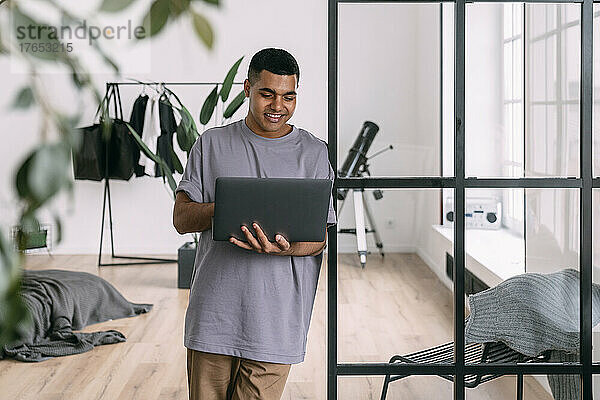 Lächelnder Mann benutzt Laptop und lehnt zu Hause an einer Glaswand