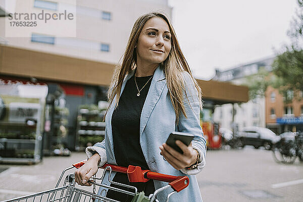 Junge Frau steht mit Smartphone und Einkaufswagen auf Fußweg