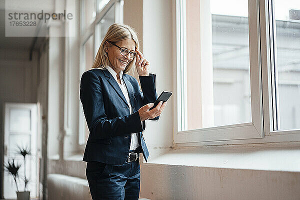 Lächelnde Geschäftsfrau benutzt Smartphone am Fenster am Arbeitsplatz
