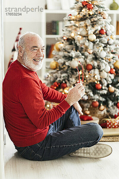 Fröhlicher älterer Mann mit Brille sitzt zu Hause mit gekreuzten Beinen vor dem Weihnachtsbaum