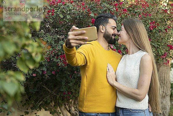 Heterosexual couple rubbing noses and taking selfie on smart phone in garden