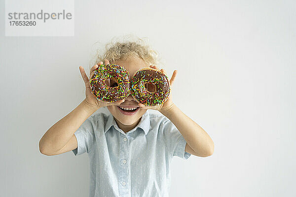 Happy girl covering eyes with doughnuts standing in front of wall