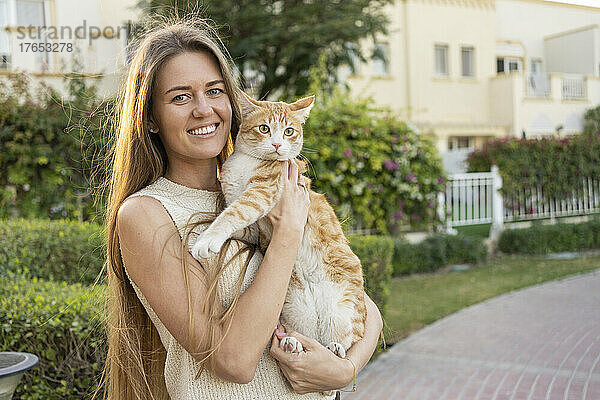 Glückliche Frau mit langen blonden Haaren hält rothaarige Katze im Garten