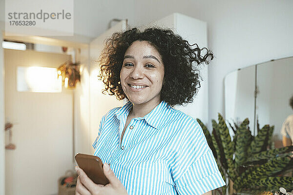 Lächelnde Frau mit Smartphone steht zu Hause im Wohnzimmer