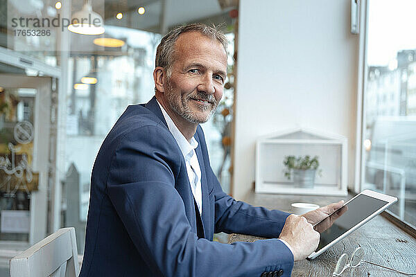 Lächelnder Geschäftsmann sitzt am Tisch am Glasfenster mit Tablet-PC