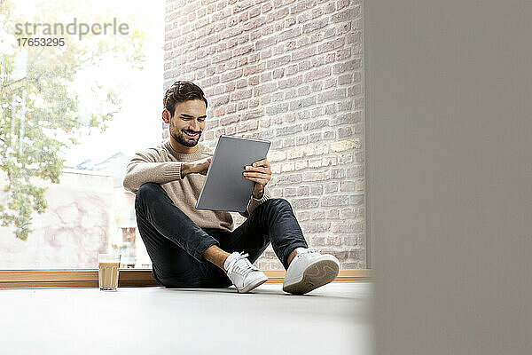 Mann sitzt mit Tablet-PC vor Glasfenster zu Hause