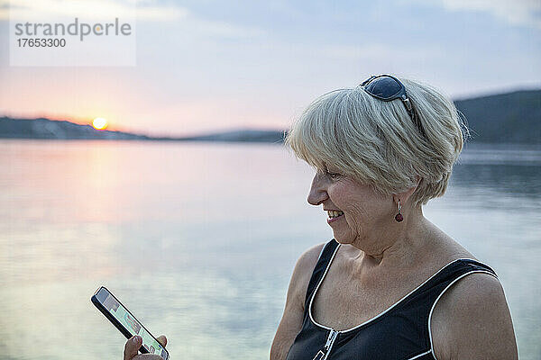 Lächelnde ältere Frau  die bei Sonnenuntergang einen Videoanruf über ihr Smartphone auf dem Seeweg tätigt