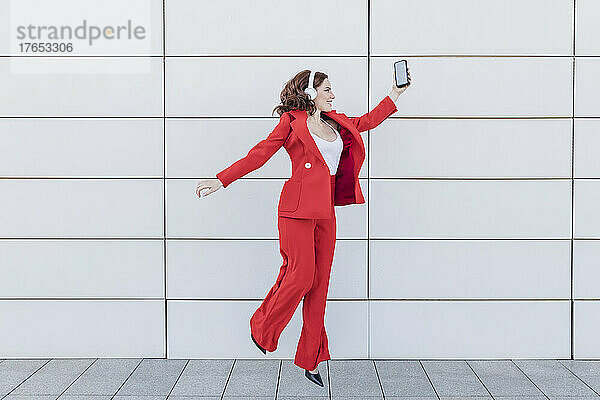 Carefree businesswoman with smart phone jumping in front of wall