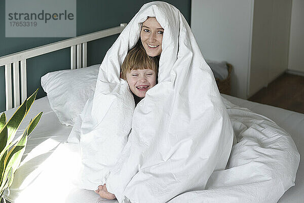 Glückliche Mutter und Sohn  in eine Decke gehüllt  sitzen zu Hause im Bett