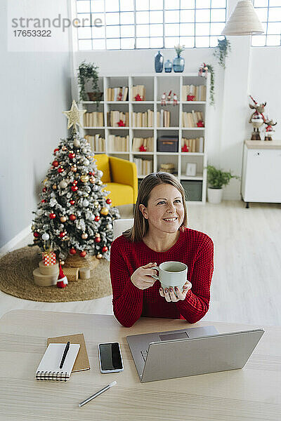 Glückliche Frau mit Kaffeetasse sitzt am Tisch im Wohnzimmer