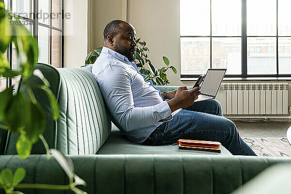 Geschäftsmann hält Laptop und Mobiltelefon in der Hand und sitzt auf dem Sofa im Büro