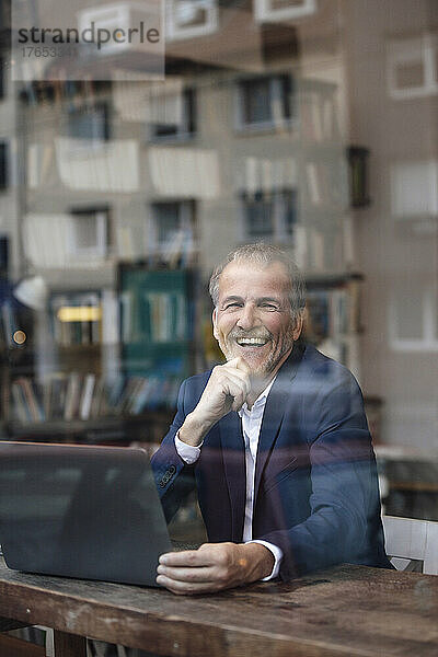 Fröhlicher Geschäftsmann mit Laptop  der durch die Glasscheibe im Café gesehen wird