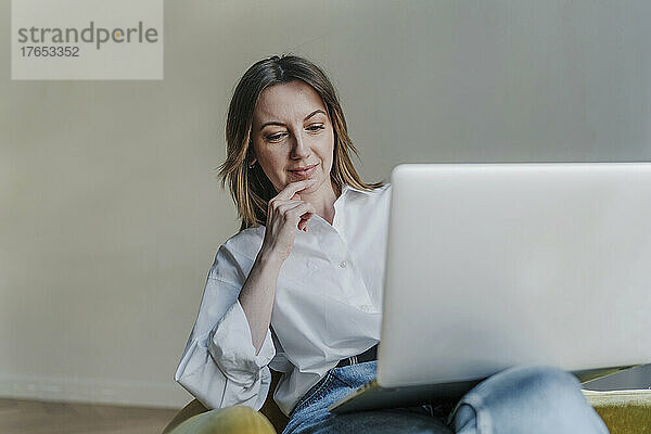 Lächelnde Frau mit Laptop sitzt auf einem Sessel vor weißem Hintergrund