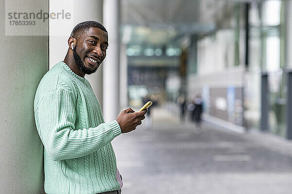 Smiling man holding mobile phone standing by wall
