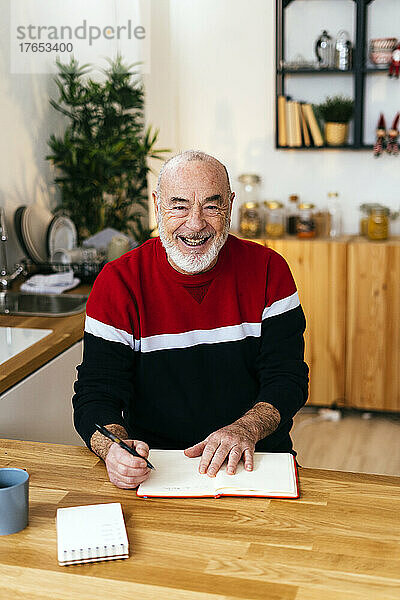 Fröhlicher älterer Mann mit Tagebuch sitzt am Tisch in der Küche
