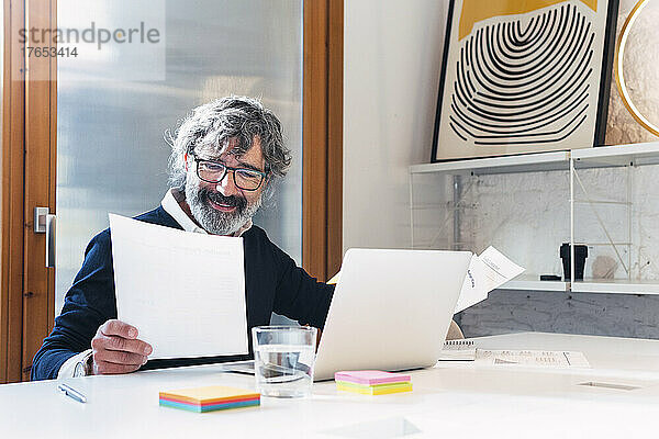 Lächelnder Freiberufler liest Dokumente und sitzt mit Laptop am Tisch