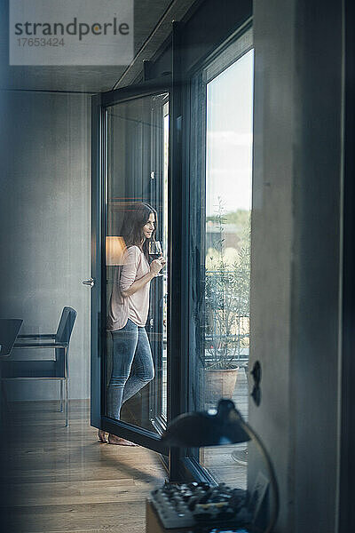 Frau mit Weinglas steht zu Hause am Fenster
