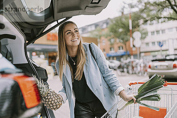 Glückliche junge Frau mit Einkaufswagen  der Lebensmittel im Kofferraum eines Autos lädt