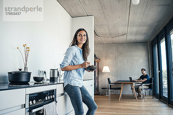 Lächelnde Frau mit Schüssel steht in der Küche mit Freiberuflerin  die zu Hause Laptop benutzt