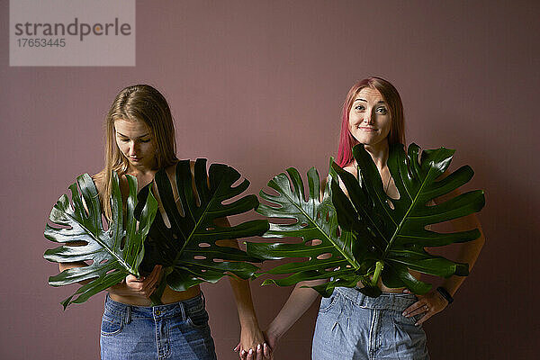 Freunde mit Monstera-Blättern halten sich an den Händen und stehen vor farbigem Hintergrund