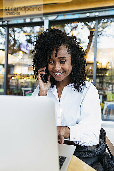 Glückliche Geschäftsfrau  die mit einem Laptop im Straßencafé sitzt und ihr Mobiltelefon in die Hand nimmt