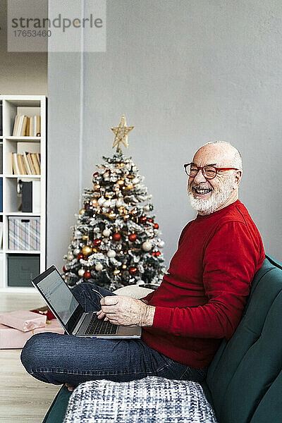 Fröhlicher älterer Mann mit Laptop sitzt zu Hause auf dem Sofa im Wohnzimmer