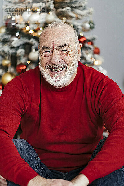 Happy senior man sitting in front of decorated Christmas tree at home