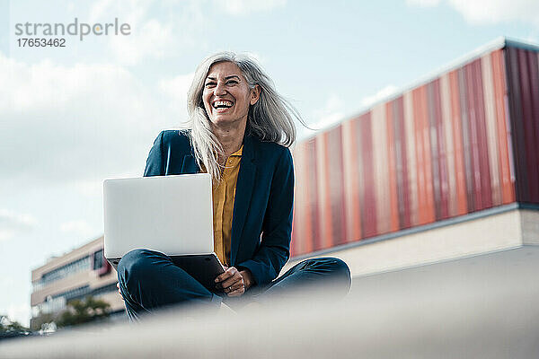 Glückliche Geschäftsfrau sitzt lachend mit Laptop im Büropark
