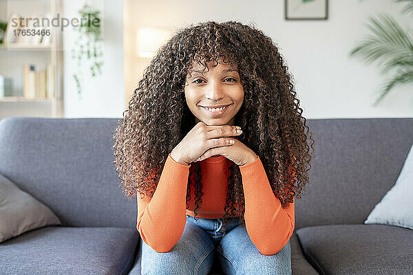 Lächelnde junge Frau mit lockigem Haar sitzt auf dem Sofa im Wohnzimmer