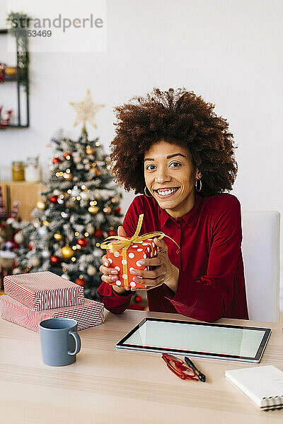 Glückliche Frau mit Weihnachtsgeschenken sitzt zu Hause mit Tablet-PC