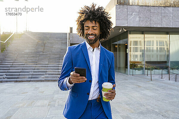 Lächelnder Geschäftsmann hält Einwegbecher in der Hand und geht mit Mobiltelefon auf Fußweg
