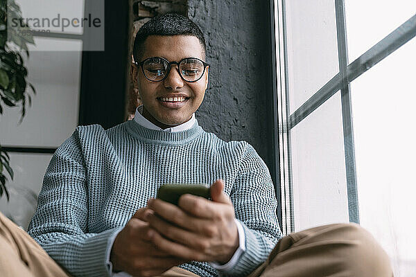 Glücklicher Mann  der zu Hause am Fenster sitzt und Textnachrichten über sein Smartphone sendet