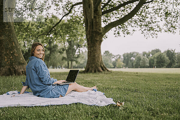 Lächelnde Frau mit Laptop sitzt auf Picknickdecke im Park