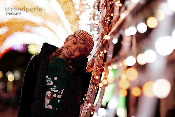 Glückliches Mädchen mit Strickmütze  das neben Weihnachtsbeleuchtung steht