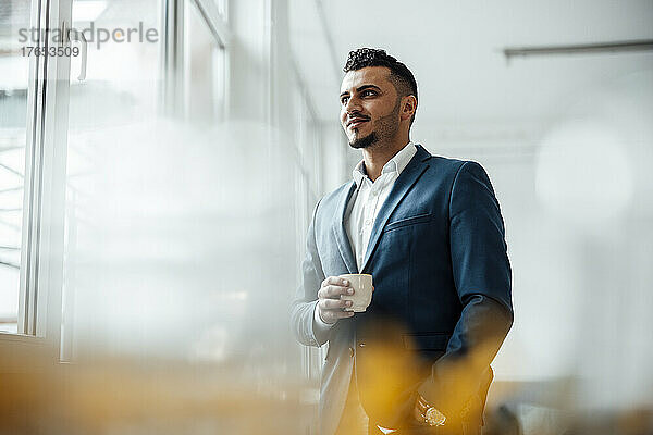 Geschäftsmann hält Kaffeetasse in der Hand und schaut durchs Fenster  während er im Büro steht