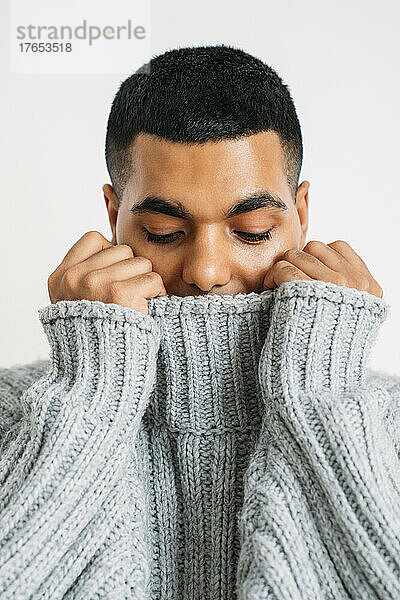 Young man covering face with gray sweater against white background