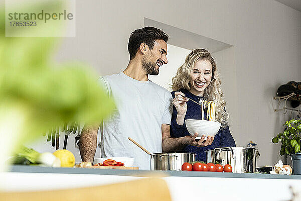 Happy man holding bowl of noodles standing with girlfriend in kitchen