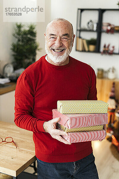 Cheerful senior man holding gift boxes standing by table at home