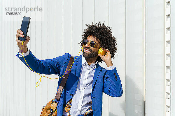 Lächelnder Geschäftsmann  der über Kopfhörer Musik hört und ein Selfie mit dem Mobiltelefon vor der Wand macht