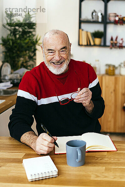 Happy senior man holding eyeglasses sitting with diary at table