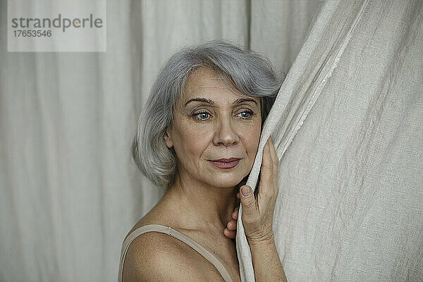 Nachdenkliche ältere Frau mit grauem Haar berührt zu Hause den Vorhang